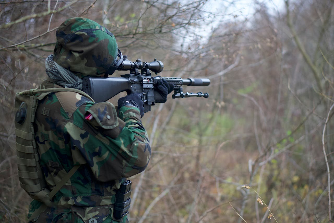 Soldier in camouflage aiming a rifle in a dense forest setting during daylight.